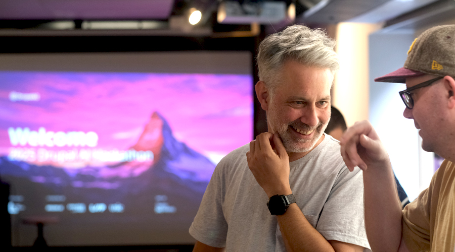 Attendees smiling and talking together at the Drupal AI Hackathon in Switzerland, with a large, glowing presentation projector screen in the background displaying a welcome message.