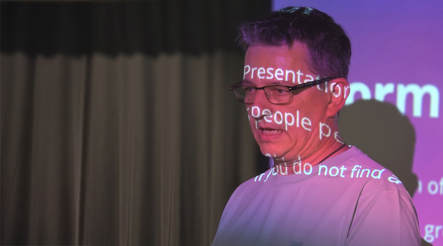 A close-up profile of a tech presenter giving a speech on stage, with the white text of his digital presentation slides illuminated and projected directly across his face and shirt.